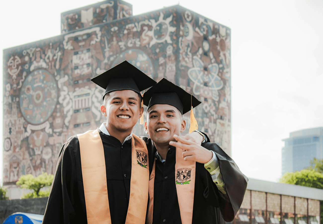 Two graduates smiling with their mortarboards and stoles in front of a historic building.